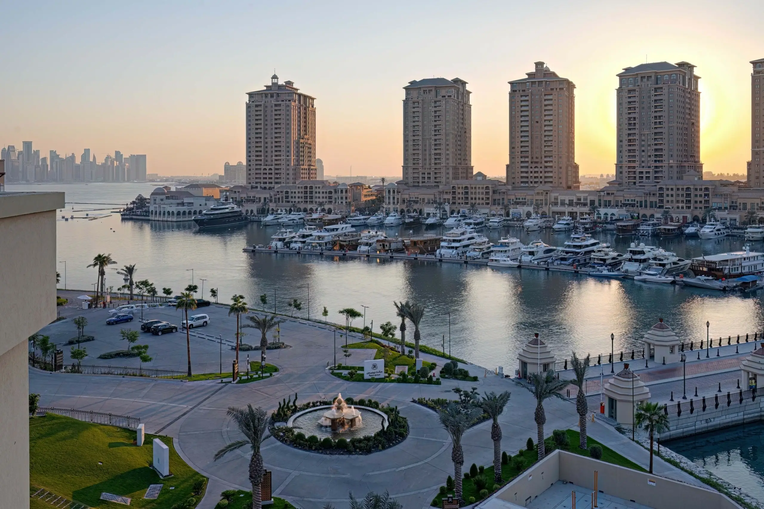 A marina with buildings, yachts, and a fountain.